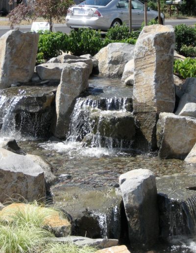 A Greenhaven Landscapes-designed water feature - a Vancouver WA backyard waterfall with large stones and shrubbery planted along it's edges