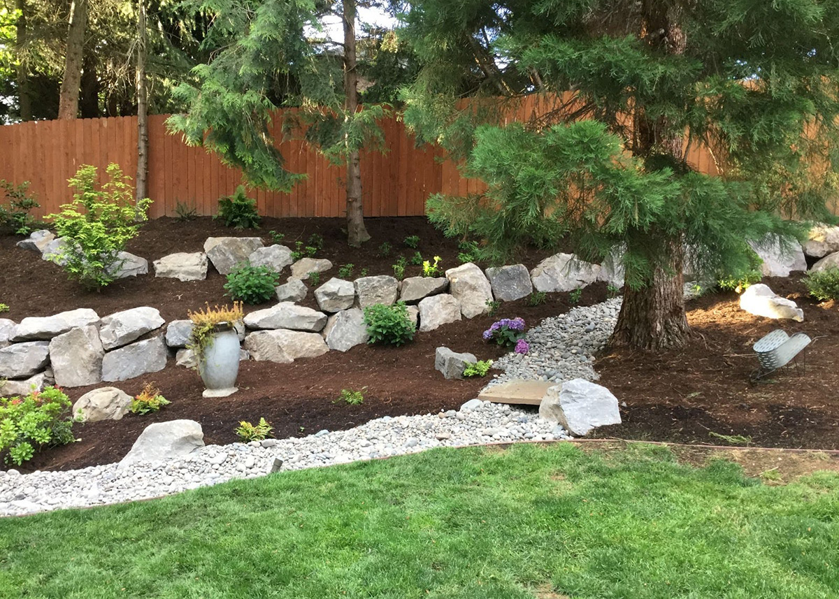 a backyard landscape design by Greenhaven Landscapes with boulder wall and rock drainage path